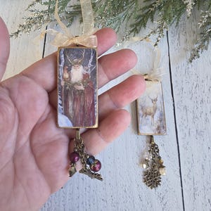 May include: Two rectangular hanging ornaments featuring printed imagery. One depicts a bearded figure with antlers in a snowy forest, the other a deer in a winter landscape. Both have gold ribbon loops, bronze-toned metal frames, and dangling charms.