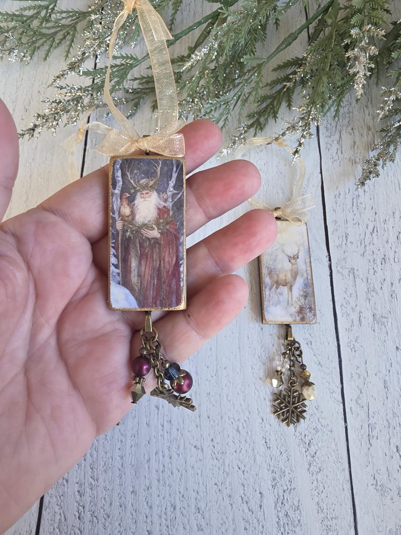 May include: Two rectangular hanging ornaments featuring printed imagery. One depicts a bearded figure with antlers in a snowy forest, the other a deer in a winter landscape. Both have gold ribbon loops, bronze-toned metal frames, and dangling charms.