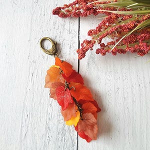 May include: A fall-themed keychain featuring a cluster of translucent, colorful leaves in shades of red, orange, and yellow. The leaves are attached to a bronze-colored clasp. Decorative red berries and green stems are in the background.