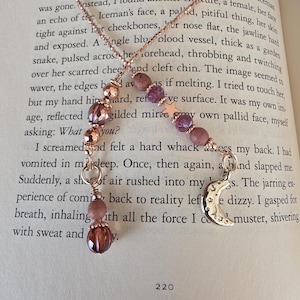 May include: A Y-shaped necklace featuring a rose gold chain, faceted purple and pink beads, a small five-pointed star charm, and a silver-toned crescent moon pendant. The necklace is draped over an open book page with visible printed text.