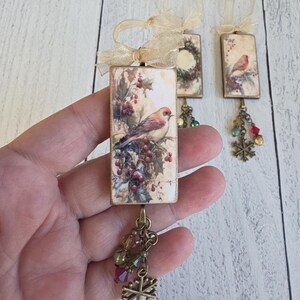 May include: Three rectangular holiday ornaments featuring vintage-style bird and wreath illustrations. Each ornament has a gold ribbon loop, a dangling bronze-toned chain with multi-colored glass beads, and a small metal snowflake charm.