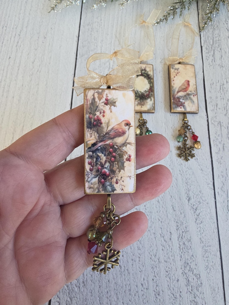 May include: Three rectangular holiday ornaments featuring vintage-style bird and wreath illustrations. Each ornament has a gold ribbon loop, a dangling bronze-toned chain with multi-colored glass beads, and a small metal snowflake charm.