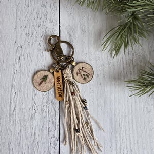May include: A keychain with a bronze clasp and key ring. It features two circular charms with embroidered designs of a tree and mountains. A small wooden tag reads "wander". The keychain has a tassel of natural-colored twine with decorative beads.