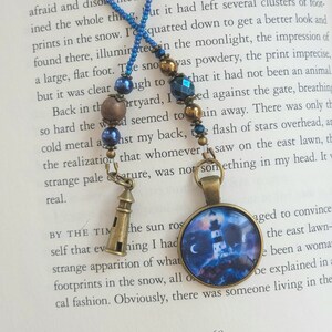 May include: Two silver tone charm necklaces with lighthouse pendants. One pendant is a small, detailed lighthouse with a blue and white design. The other pendant is a round, glass charm with a blue and white lighthouse image and a crescent moon. The necklaces are on a book with the page number 168 visible.