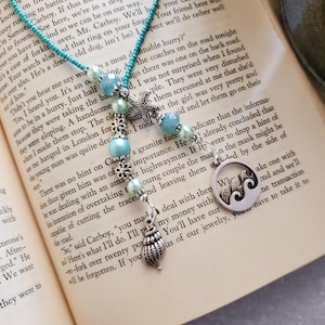 May include: A beaded necklace featuring a turquoise cord, light blue and mint green beads, and silver-toned metal charms. The charms include a starfish, a circular pendant with a wave design, and a seashell, all arranged on a book page.