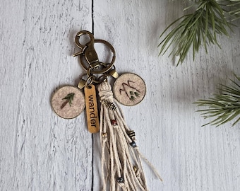 May include: A keychain featuring a bronze lobster clasp, two small circular embroidered patches with tree motifs, a rectangular wooden tag engraved with the word wander, and a long beige cord tassel adorned with small metallic and colored beads.