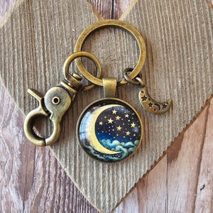 May include: A bronze-toned keychain featuring a circular pendant with a glass cabochon displaying a yellow crescent moon, golden stars, and blue clouds against a dark night sky. The keychain includes a lobster clasp and a small crescent moon charm.