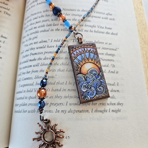 May include: A bookmark featuring a rectangular pendant with a sun and ocean wave illustration, attached to a beaded chain of blue, orange, and metallic beads. The chain ends with a copper-toned sun charm, creating a decorative accessory.