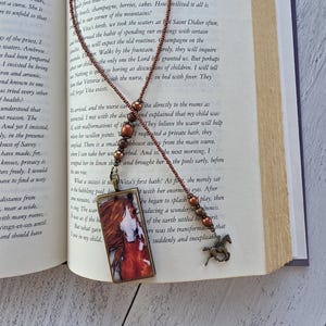 May include: A bookmark featuring a rectangular pendant with a horse illustration, attached to a beaded chain made of small brown beads and larger metallic copper-toned spheres, ending with a small bronze-colored horse charm at the bottom.