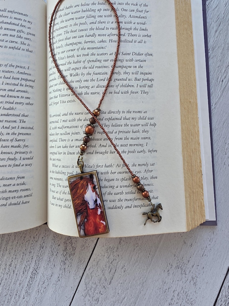 May include: A bookmark featuring a rectangular pendant with a horse illustration, attached to a beaded chain made of small brown beads and larger metallic copper-toned spheres, ending with a small bronze-colored horse charm at the bottom.
