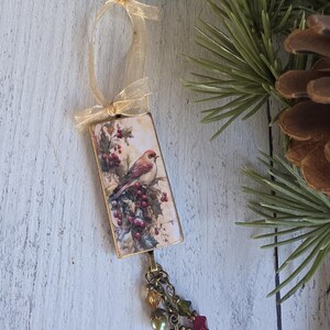 May include: A rectangular hanging ornament featuring a printed image of a bird perched on holly branches with red berries. The ornament is topped with a gold ribbon bow and finished at the bottom with a bronze chain, colorful beads, and a snowflake charm.