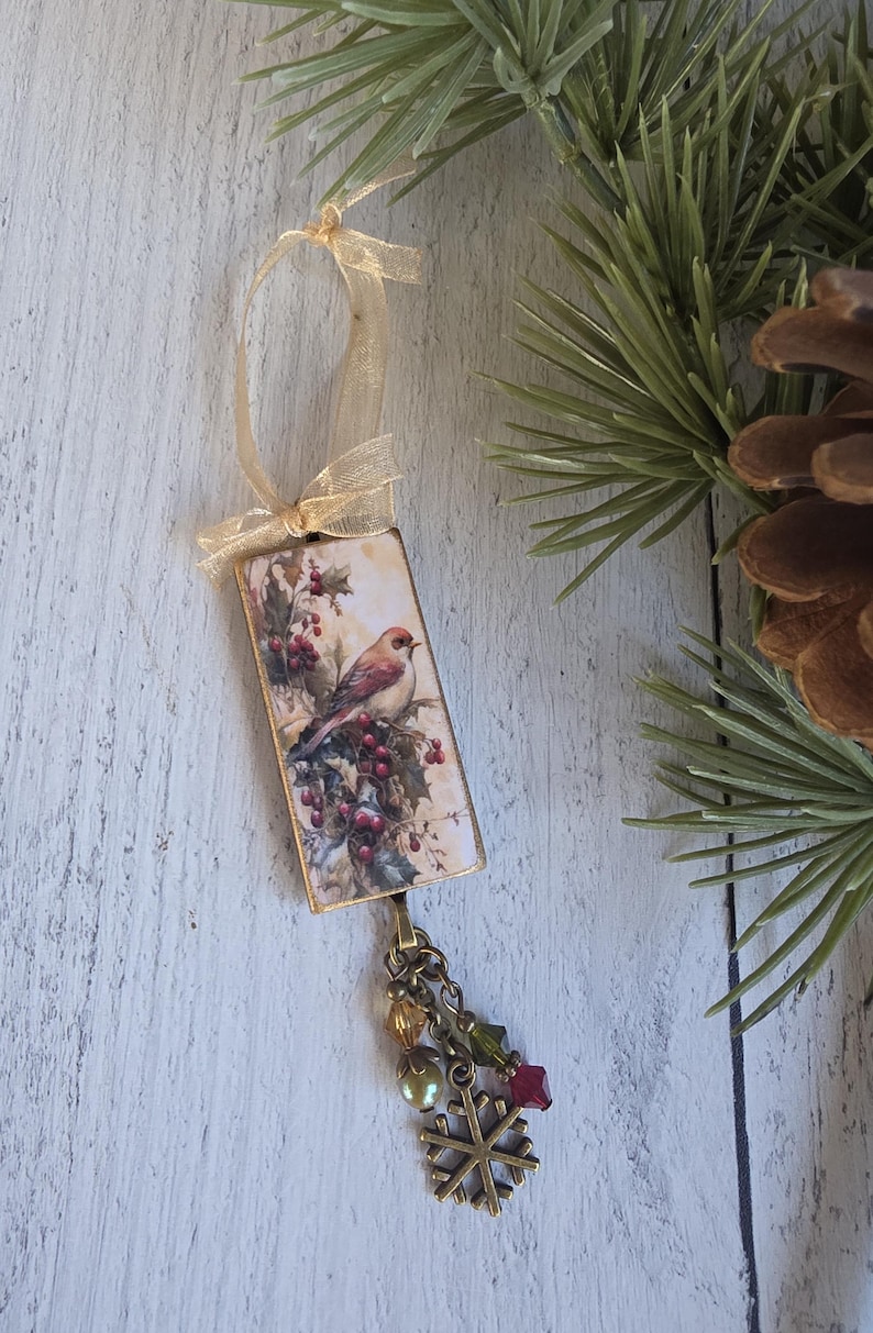 May include: A rectangular hanging ornament featuring a printed image of a bird perched on holly branches with red berries. The ornament is topped with a gold ribbon bow and finished at the bottom with a bronze chain, colorful beads, and a snowflake charm.