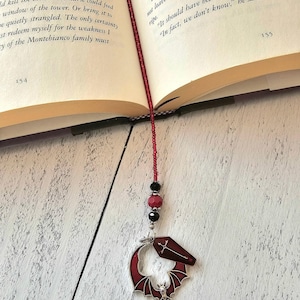 May include: A red and silver dragon-themed bookmark with a beaded strand. The bookmark features a red dragon design with a coffin and bat charm, and is placed in an open book. The beads are black and red.
