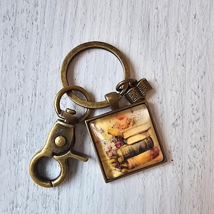 May include: A bronze-toned metal keychain featuring a square pendant with a glass cabochon display of a stack of vintage books and a small book-shaped charm. The hardware includes a circular split ring and a lobster claw clasp for attachment.