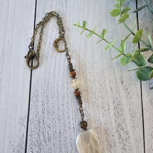 May include: A decorative hanging ornament featuring a bronze-toned metal chain with a lobster clasp and a crescent moon charm. The chain is accented with two brown wooden beads, a translucent yellow bead, and a large, smooth, off-white stone pendant.