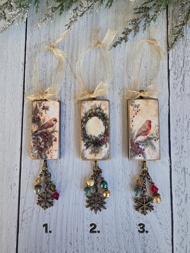May include: Three rectangular holiday ornaments featuring vintage-style bird and wreath illustrations. Each ornament has a gold ribbon loop, a bronze-toned metal frame, and a dangling charm cluster with colorful beads and a metal snowflake pendant.