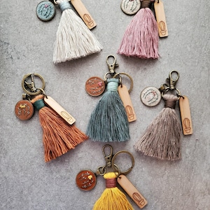 May include: Six keychains featuring colourful fabric tassels, bronze-toned metal lobster clasps, small circular embroidered patches with botanical designs, and rectangular wooden tags engraved with a simple cactus illustration.