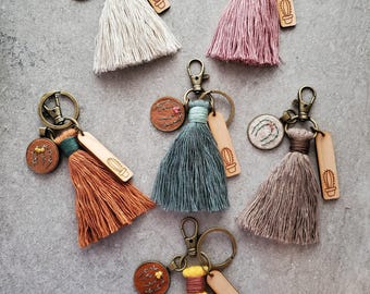 May include: Six keychains featuring colorful fabric tassels, bronze-toned metal lobster clasps, small circular embroidered patches with botanical designs, and rectangular wooden tags engraved with a simple cactus illustration.