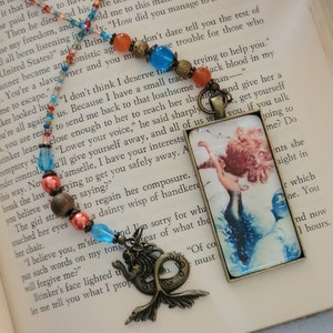 May include: A book bookmark featuring a mermaid with a blue tail and a bronze mermaid charm. The bookmark is made of a clear plastic with a bronze frame. The book is open to a page with text about a slave.
