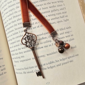 May include: A bookmark featuring a burnt orange velvet ribbon attached to an antique bronze-colored metal key charm with intricate filigree detailing. A secondary ribbon strand is adorned with two round beads in shades of brown and copper.