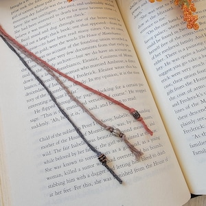 May include: Three handmade bookmarks made of twisted cord in grey, dusty pink, and light brown. Each bookmark features small metallic beads and a clear glass bead accent. The bookmarks are placed vertically across an open book page with text.
