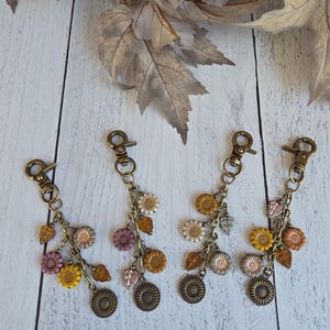 May include: Four decorative keychains featuring bronze-toned lobster clasps and chains adorned with small, multi-colored flower and leaf-shaped charms in shades of yellow, purple, orange, white, and grey, finished with a round metal sunflower charm.
