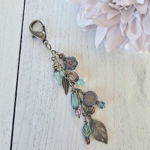 May include: A bronze-toned metal keychain featuring a lobster clasp and a cluster of dangling charms. The charms include blue and purple flower-shaped beads, translucent teardrop beads, and detailed metal leaf pendants attached to a chain.
