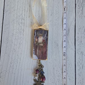 May include: A rectangular hanging ornament featuring a bearded figure in a red robe with antlers, topped with a gold ribbon. A bronze-toned charm dangles below with purple beads and a snowflake. A measuring tape shows a length of 6.5 inches.