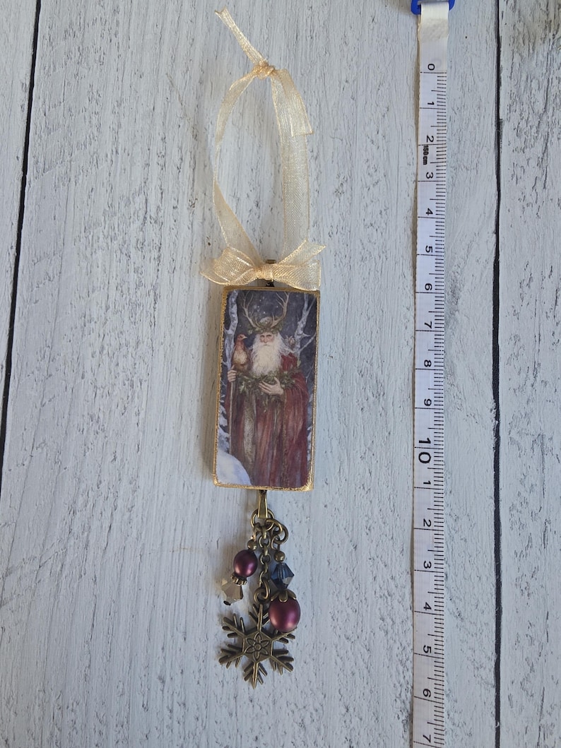 May include: A rectangular hanging ornament featuring a bearded figure in a red robe with antlers, topped with a gold ribbon. A bronze-toned charm dangles below with purple beads and a snowflake. A measuring tape shows a length of 6.5 inches.