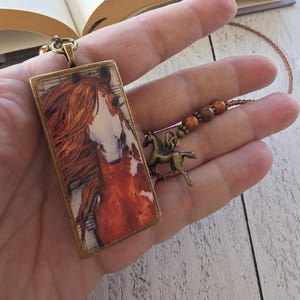 May include: A rectangular pendant featuring a detailed image of a chestnut horse with a white blaze on its face. The pendant has an antique bronze-toned metal frame and is attached to a chain adorned with small copper-colored beads and a horse charm.