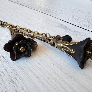 May include: A black and gold necklace with two flower pendants. The flowers are made of black polymer clay and have a vintage style. The necklace is made of brass chain and has a delicate design.