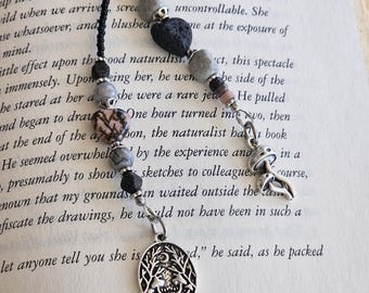 May include: A black cord bookmark featuring two strands adorned with various beads, including gray, black, and pink stone hearts. One strand ends with a silver-toned mushroom charm, while the other features a round silver pendant depicting a forest scene.