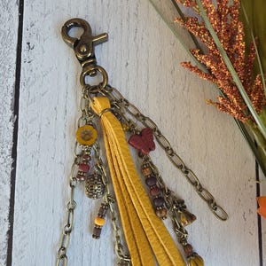 May include: A keychain featuring a brass lobster clasp and multiple hanging chains. It includes a yellow leather tassel, a red butterfly charm, a yellow flower bead, a rectangular metal charm, and various brown and yellow spherical beads.