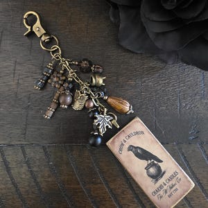 May include: A keychain featuring a bronze lobster clasp and a collection of charms including an owl, a cauldron, a leaf, and various brown and black beads. Attached is a rectangular wooden tag with a crow and cauldron illustration and text.