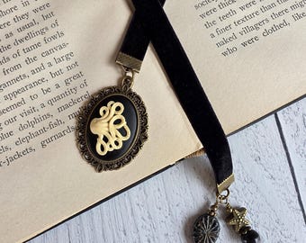 May include: A black velvet ribbon bookmark featuring an oval bronze-toned cameo pendant with a cream-colored octopus relief. The bookmark includes a dangling charm cluster with a small bronze starfish, a dark textured bead, and a smooth dark bead.
