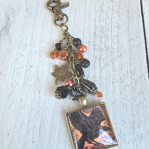 May include: A bronze-toned keychain featuring a square pendant with a black raven illustration on an orange background. The chain is adorned with various charms, including a metal maple leaf, a small black raven, and assorted orange and black beads.