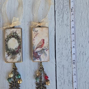 May include: Two rectangular hanging ornaments with gold ribbon loops. One features a wreath, the other a bird on a branch. Both have bronze-toned metal snowflake charms and multi-colored beads attached at the bottom. A measuring tape shows a length of 5.5 inches.