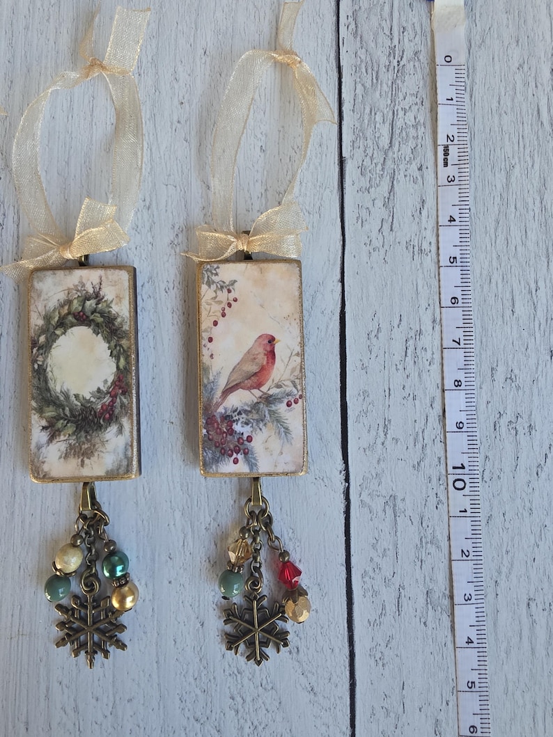May include: Two rectangular hanging ornaments with gold ribbon loops. One features a wreath, the other a bird on a branch. Both have bronze-toned metal snowflake charms and multi-colored beads attached at the bottom. A measuring tape shows a length of 5.5 inches.