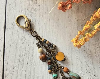 May include: A bronze-toned metal keychain featuring a lobster clasp attached to a chain with multiple dangling charms. The charms include various beads in shades of turquoise, brown, and yellow, along with small metal flower, leaf, and circular coin-shaped pendants.
