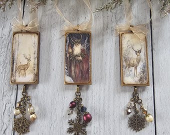May include: Three rectangular hanging ornaments featuring vintage-style illustrations of a deer, a bearded figure in a robe, and another deer. Each piece has a gold-toned metal frame, a sheer ribbon loop, and a dangling charm with beads and a snowflake.
