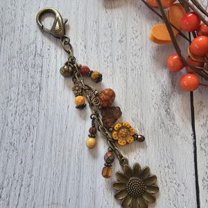 May include: A bronze-toned metal keychain featuring a lobster clasp and a multi-strand chain design. The accessory is adorned with various beads in shades of orange, brown, and yellow, along with charms shaped like leaves and flowers.