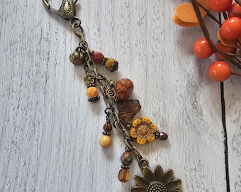 May include: A bronze-toned metal keychain featuring a lobster clasp and a multi-strand chain design. The accessory is adorned with various beads in shades of orange, brown, and yellow, along with charms shaped like leaves and flowers.