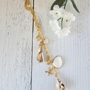 May include: A gold-coloured keychain with a lobster clasp and chain, decorated with seashell charms. The charms include a starfish, scallop shells, and a conch shell. White flowers are in the background.