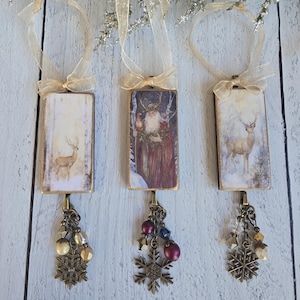 May include: Three rectangular hanging ornaments with gold ribbon loops. Each features a winter-themed image, including a deer in a snowy forest and a bearded figure in a red cloak. Each ornament has a bronze-toned metal snowflake charm and beads.