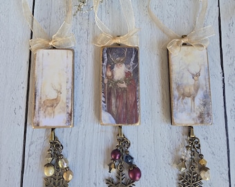 May include: Three rectangular hanging ornaments with gold ribbon loops. Each features a winter-themed image, including a deer in a snowy forest and a bearded figure in a red cloak. Each ornament has a bronze-toned metal snowflake charm and beads.