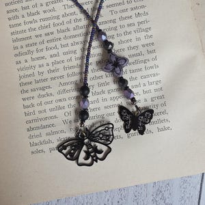 May include: A beaded bookmark featuring a long strand of small purple and black beads. The strand ends with a larger purple flower-shaped bead, a small black butterfly charm, and a larger open-work metal moth charm with moon motifs.