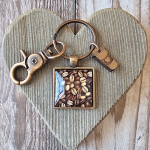 May include: A bronze-colored keychain with a square charm featuring a close-up image of coffee beans. The keychain also includes a small charm shaped like a coffee cup with a heart on it.