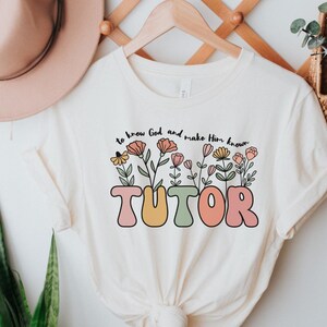 May include: A white t-shirt with a floral design and the word "TUTOR" in colorful, bubble letters. The text above the word "TUTOR" reads "to know God and make him known."