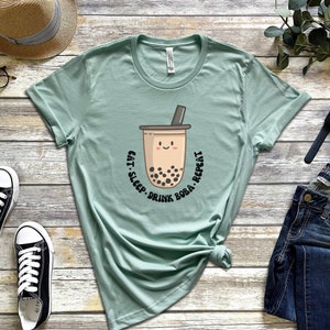 May include: A light green t-shirt with a graphic of a boba tea cup with a smiling face and the text "Eat. Sleep. Drink. Boba. Repeat."