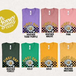 May include: A set of ten t-shirts in various colors featuring a graphic of a smiling flower with the text "First Day" in a checkered background. The t-shirts are available in Heather Team Purple, Heather Mauve, Pink, Heather Forest, Kelly, Gold, and Heather Mustard.